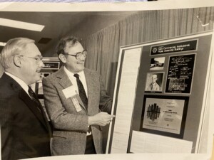 A black and white photo of two men in suits look at a poster board display together focused on energy efficiency in commercial buildings. 